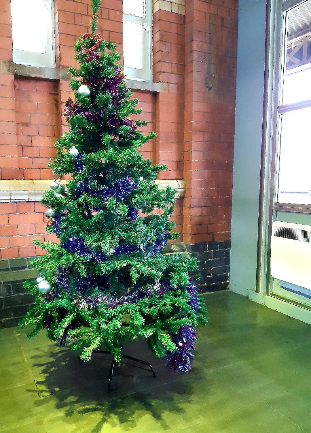 A Christmas tree standing in front of a brick wall in a station waiting room, decorated with tinsel and silver baubles.
