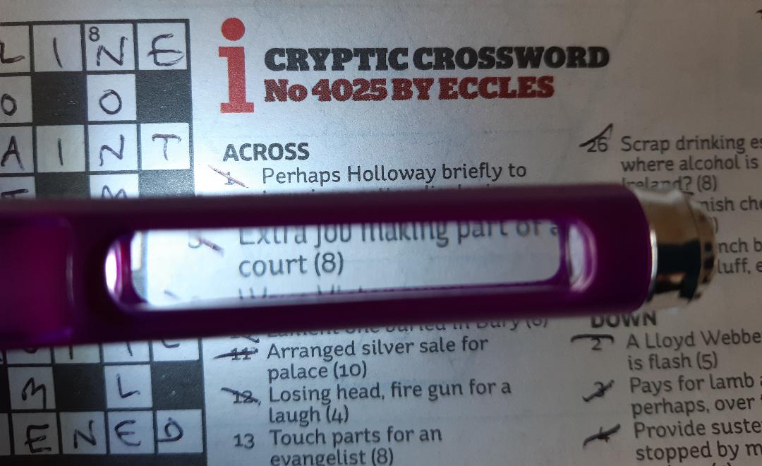 The end of a purple pen held over a cryptic crossword in a newspaper, demonstrating the small magnifying panel and bright light in the side of the pen, making it easier to read the crossword clues.